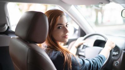 A woman looks over her shoulder towards the back seat while sitting at the wheel of a stationary car with a serious look on her face.