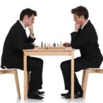 two men in business suits sit across from each other at a table with a chess board on it. Both hold their hands to their chins and look down in serious contemplation of their next move.
