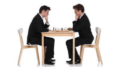 two men in business suits sit across from each other at a table with a chess board on it. Both hold their hands to their chins and look down in serious contemplation of their next move.