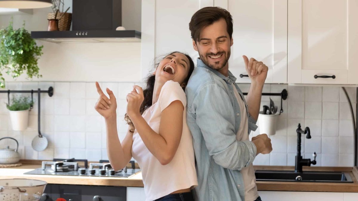 Man and woman dance back to back in kitchen.