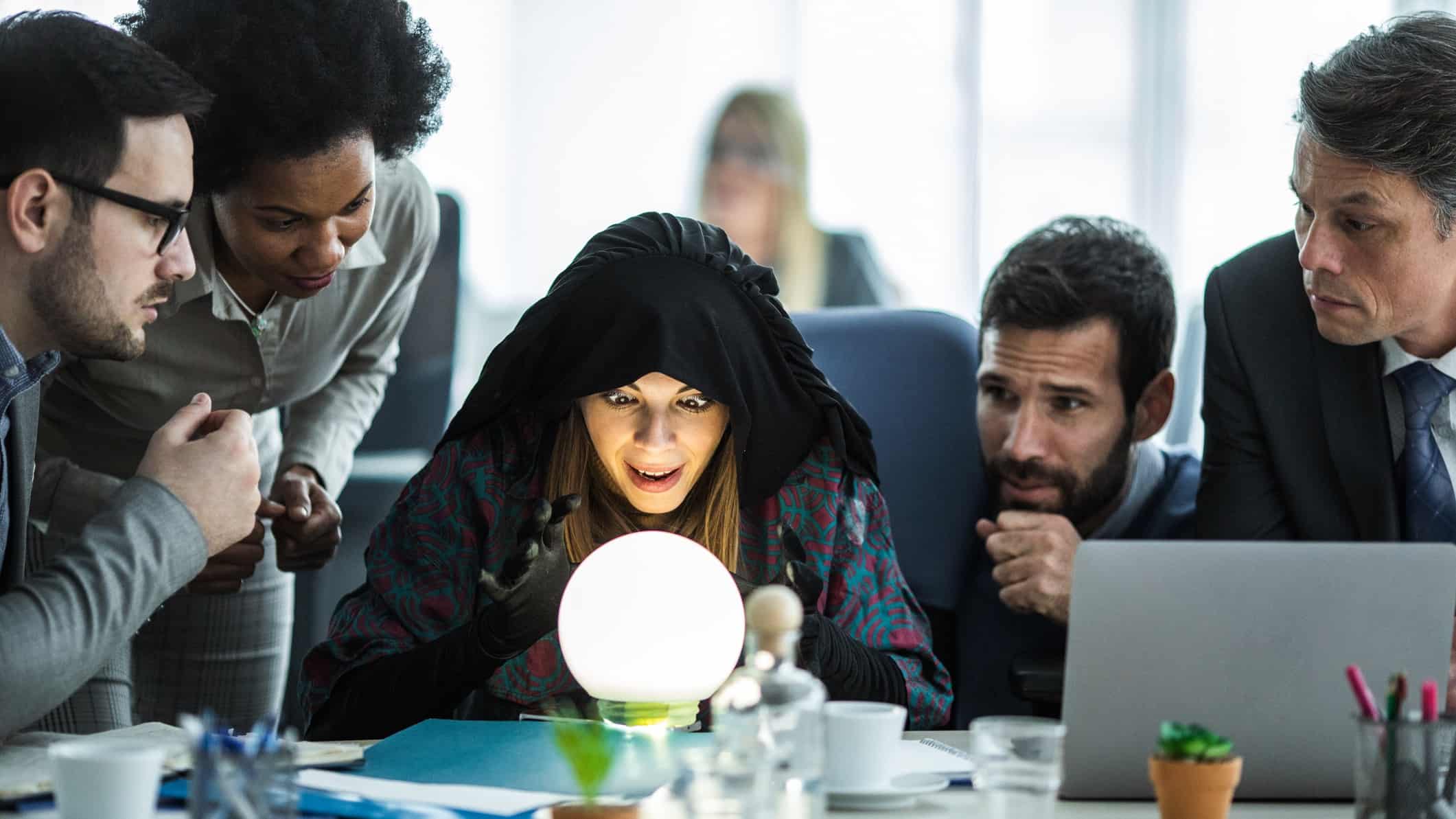 A fortune teller looks into a crystal ball in an office surrounded by business people.