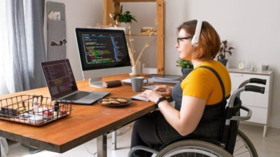 a woman in a wheelchair sits at her desk in her home with headphones on and looking at a computer screen of figures. monitoring the CBA share price