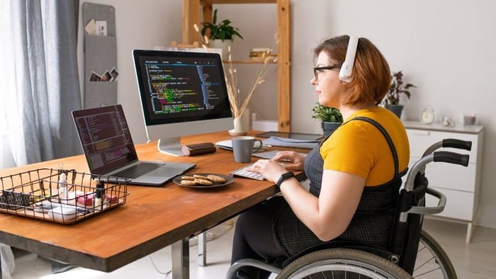 a woman in a wheelchair sits at her desk in her home with headphones on and looking at a computer screen of figures.