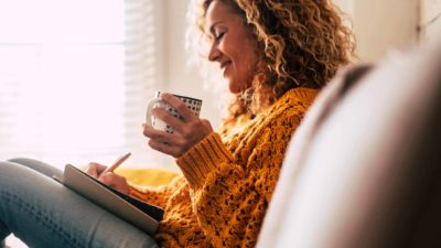 A woman in yellow jump holds a coffee and writes in a diary.