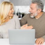 A man and woman sit next to each other looking at each other and feeling excited and surprised after reading good news about their shares on a laptop.
