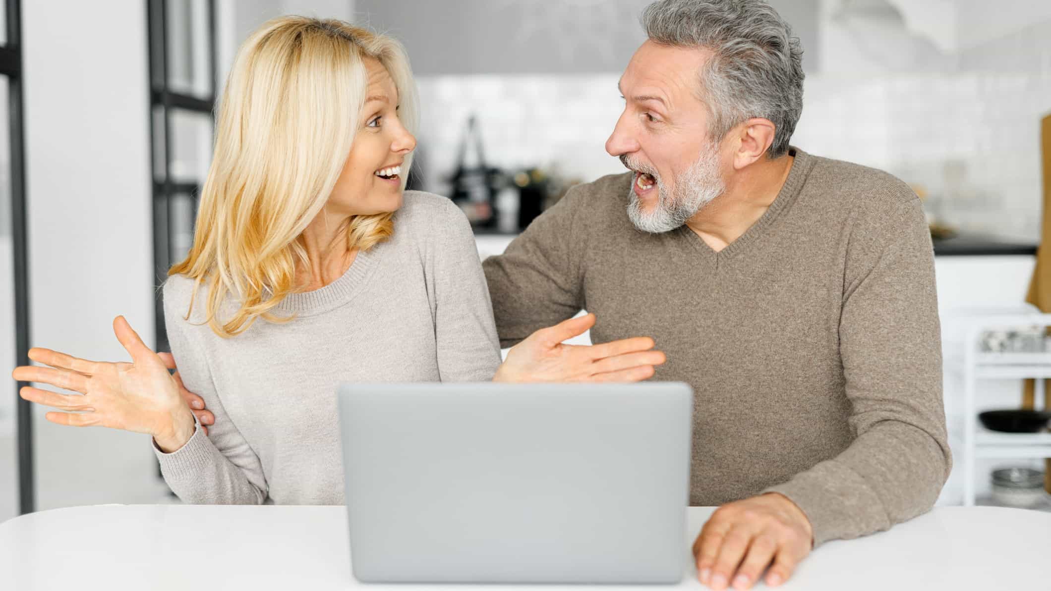 A man and woman sit next to each other looking at each other and feeling excited and surprised after reading good news about their shares on a laptop in front of them