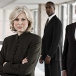 A panel of formidable business people stand in a group with serious looks on their faces as if in judgement of what's before them.