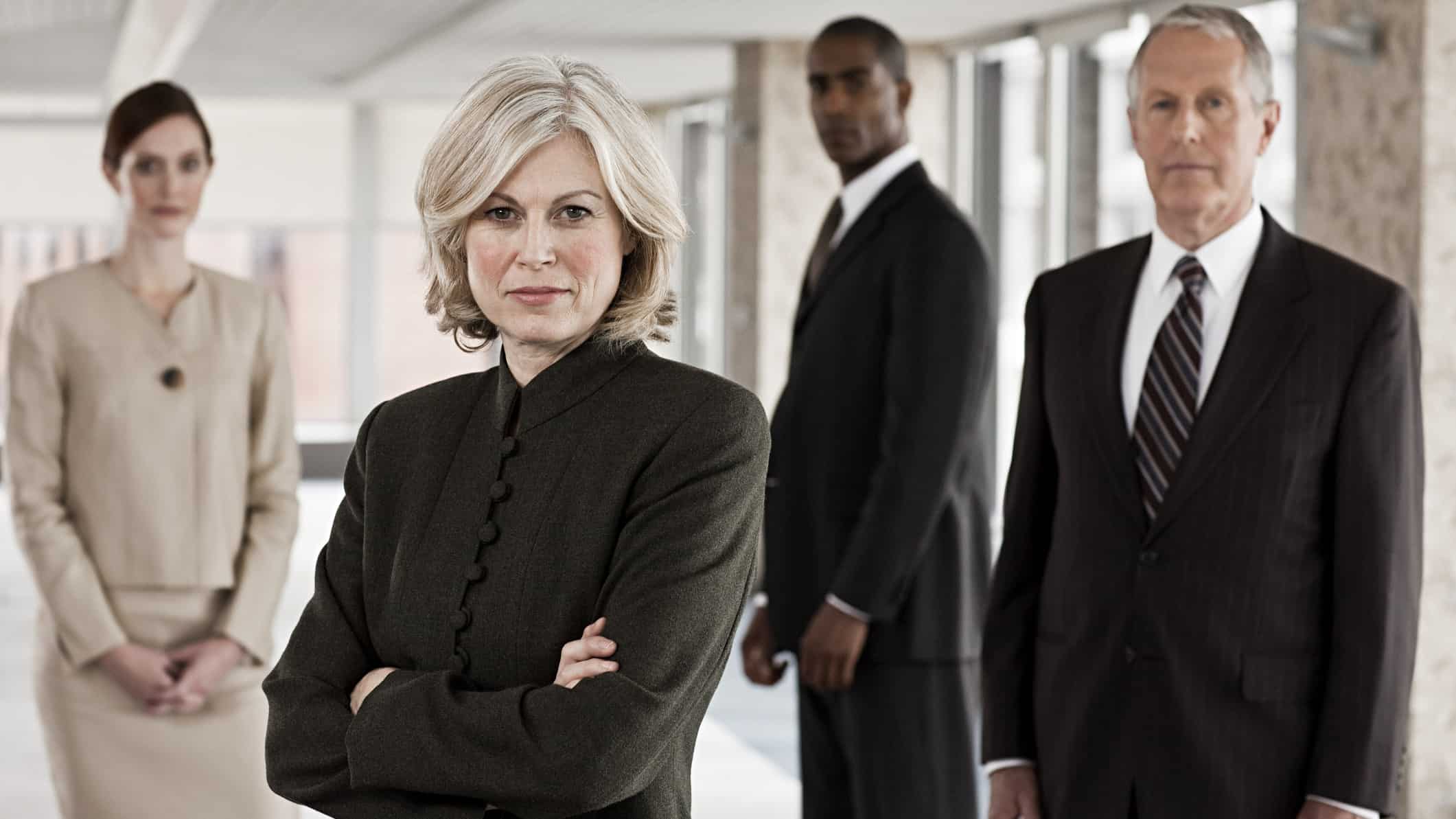 A panel of formidable business people stand in a group with serious looks on their faces as if in judgement of what's before them.