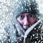 A man peers out from a high collared jacket with just his eyes and nose visible amid a swirling snowstorm.