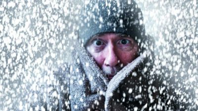 A man peers out from a high collared jacket with just his eyes and nose visible amid a swirling snowstorm.