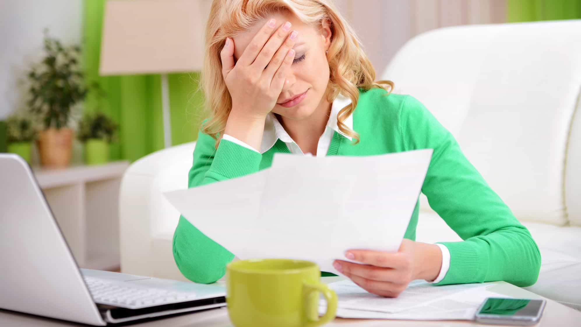 a woman wearing green and sitting in a green room with a green coffee cup puts her hand to her forehead in dismay while looking at papers sitting at her computer.