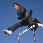 A man in a business suit jumps over a hurdle with a blue sky background.