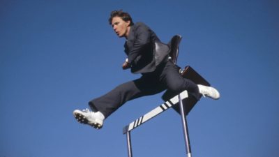 a man in a business suit jumps over a hurdle with a blue sky background.