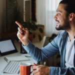 An investor looks happy holding a finger to his computer screen while holding a coffee cup in a home office scenario.