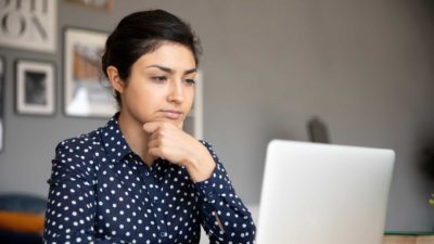 A young woman sits at her desk in deep contemplation with her hand to her chin while seriously considering information she is reading on her laptop