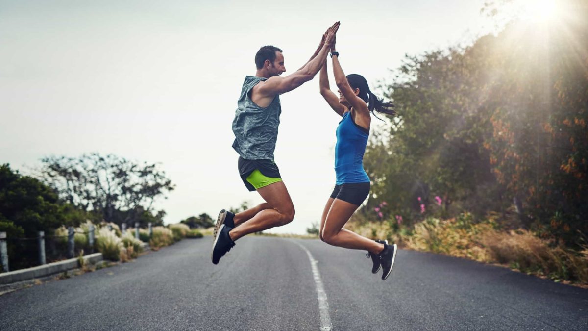 A man and woman jump in the air and high five with both hands on a road after running.