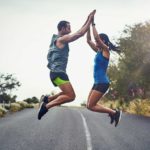A man and woman jump in the air and high five with both hands on a road after running.
