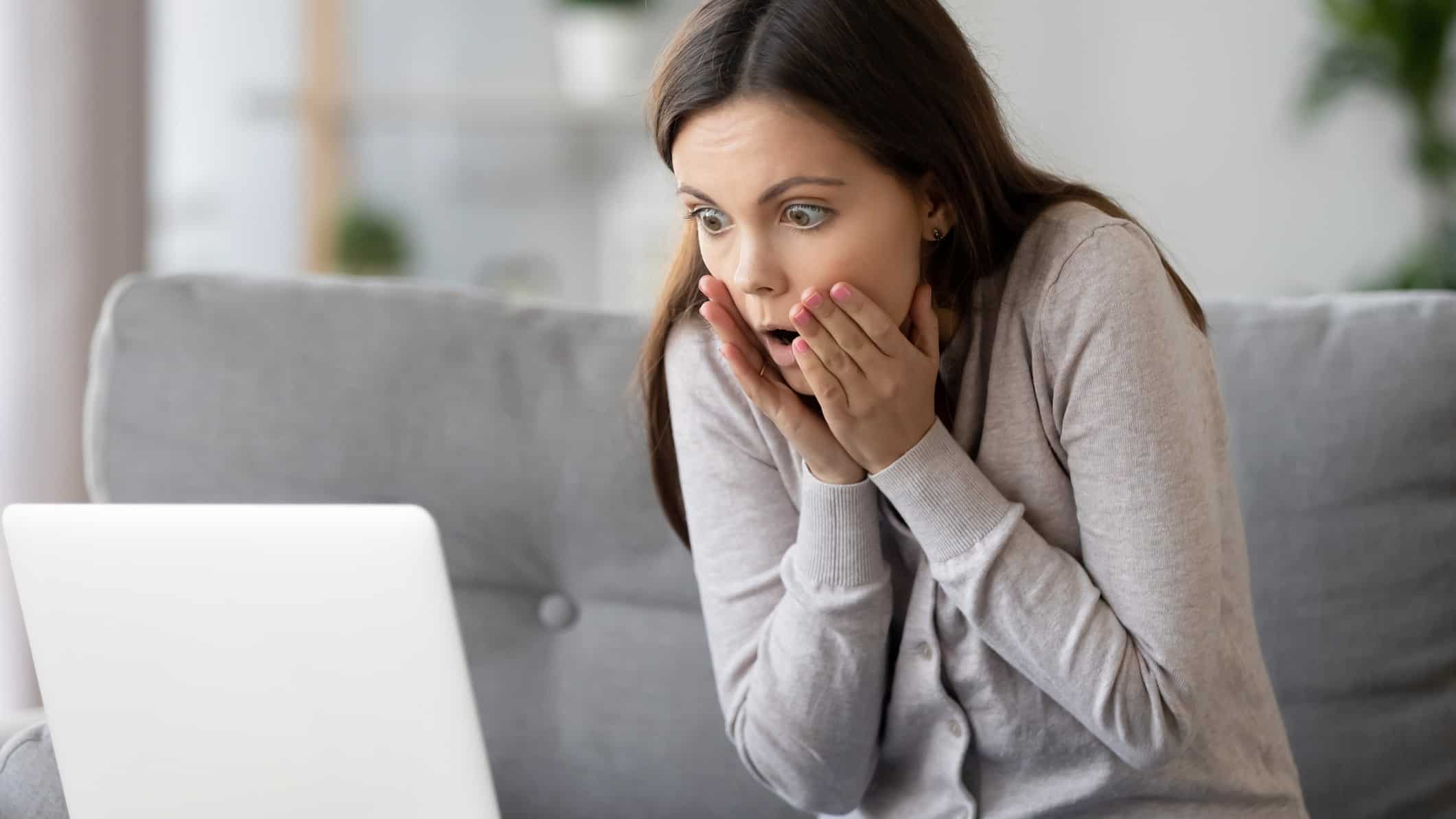 A woman holds her hands to the side of her face as she sits back in shock at something she is reading or seeing on her computer screen.