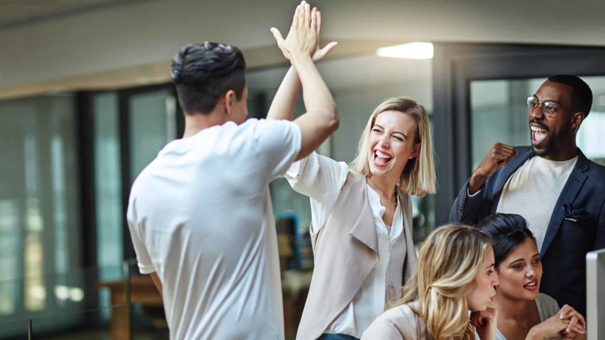 a group of office workers high five each other as they celebrate good news with a couple of workers sitting at theircomuter looking into the screen.