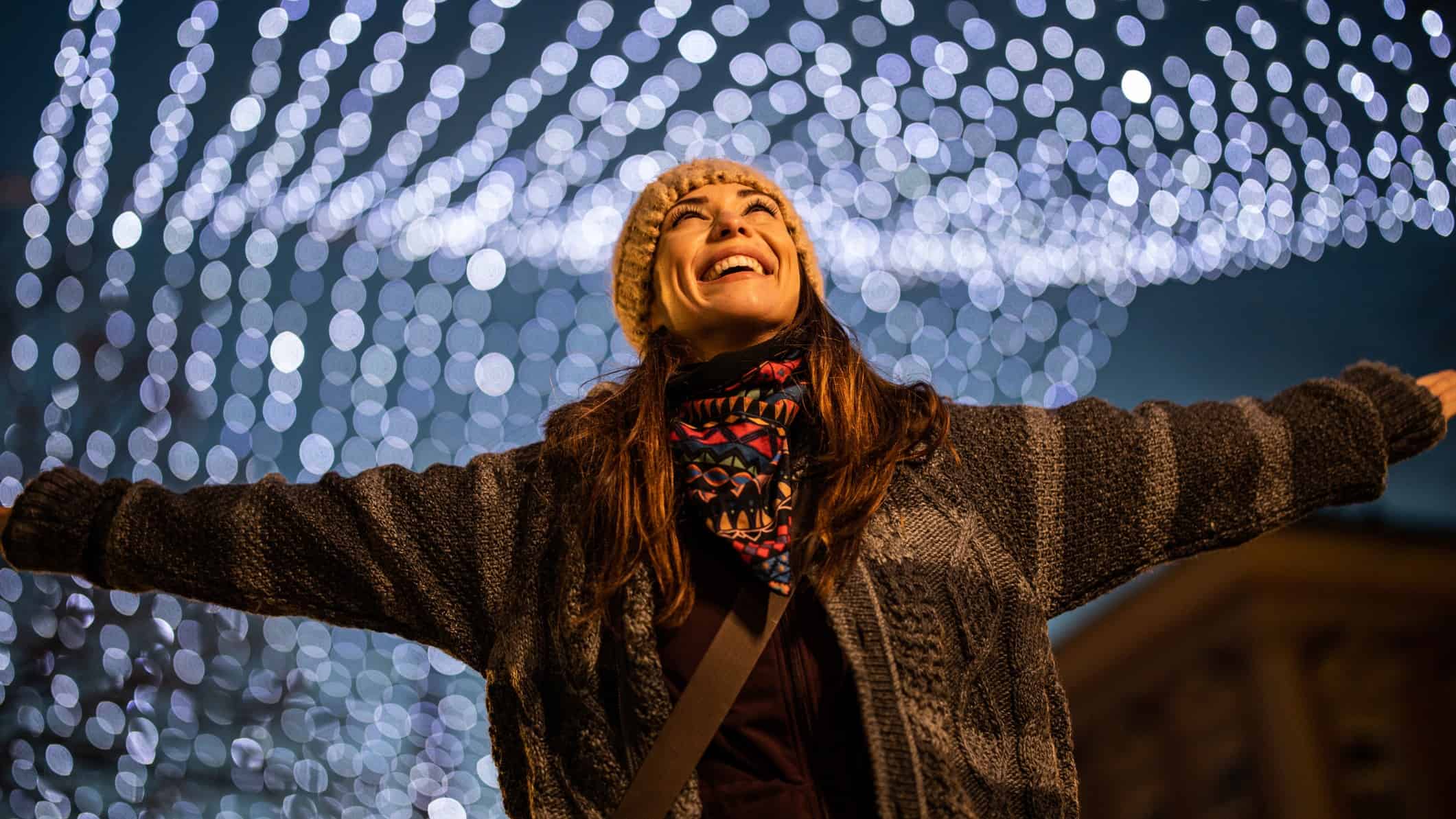 a woman holds her arms wide and looks up with an excited and joyous expression on her face as she looks up into a ceiling filled with multiple strings of glowing lights at night time.