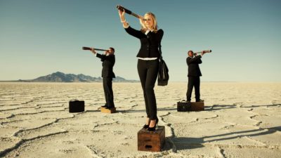 Three business people stand on platforms in the desert and look out through telescopes.