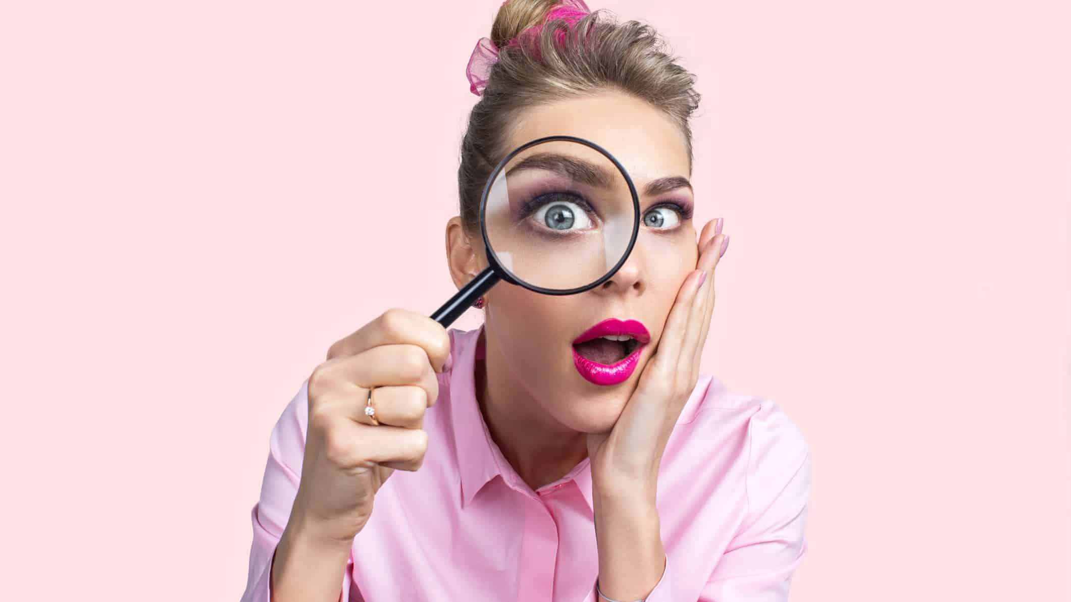 A female ASX investor looks through a magnifying glass that enlarges her eye and holds her hand to her face with her mouth open as if looking at something of great interest or surprise.