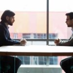 two business men sit across from each other at a negotiating table. with a large window in the background.
