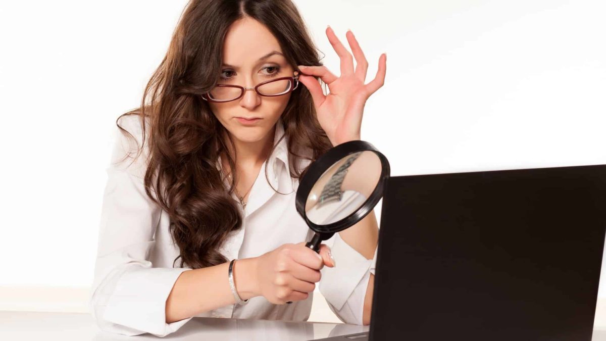 A woman with a magnifying glass adjusts her glasses as she holds the glass to her computer screen and peers closely at it.