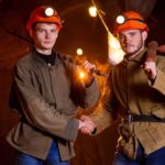 Two young male miners wearing red hardhats stand inside a mine and shake hands