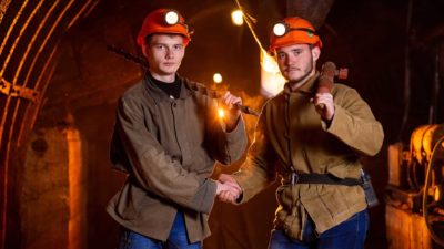 Two young male miners wearing red hardhats stand inside a mine and shake hands