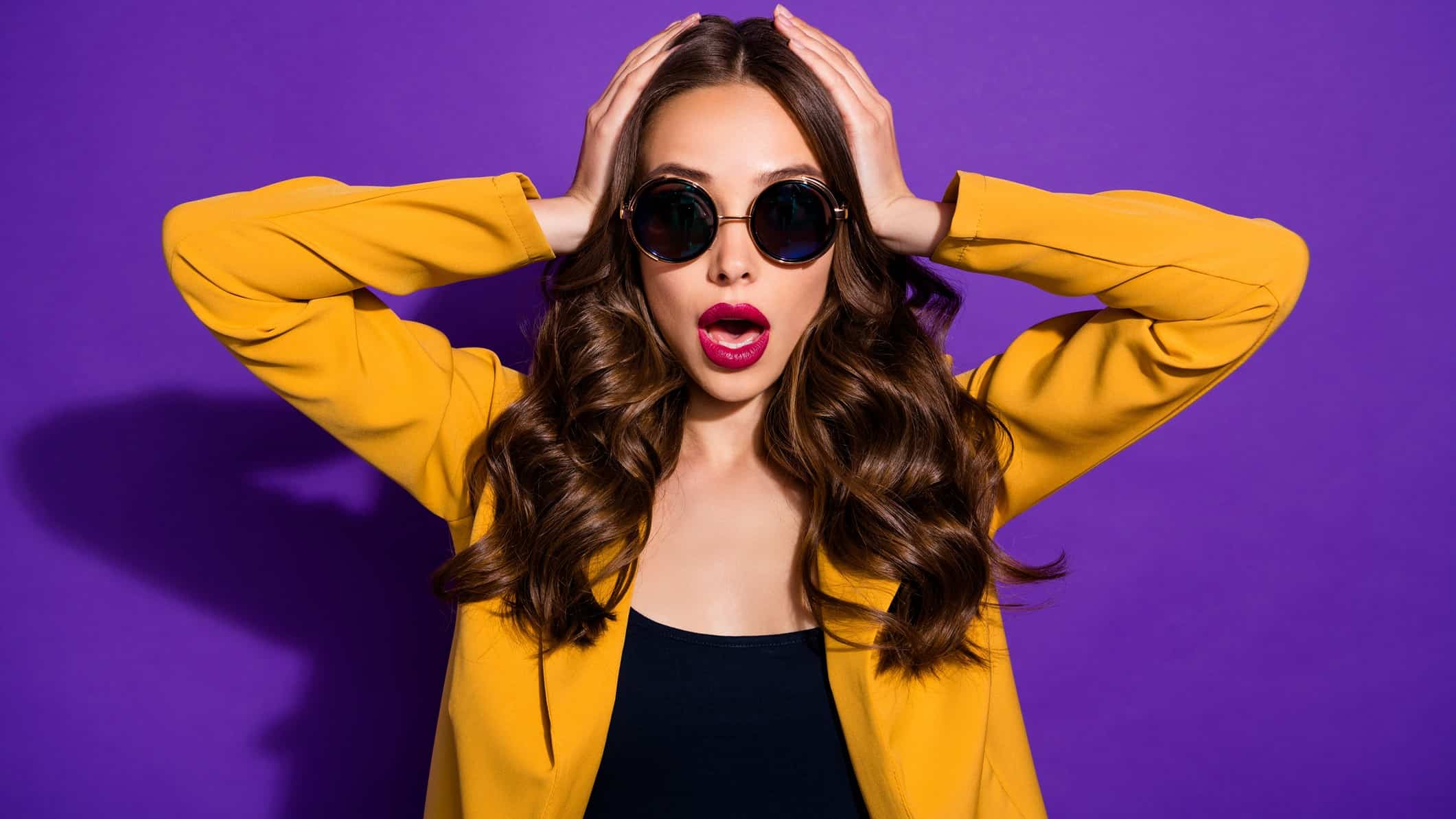 an attractive model-like woman holds her hands to her head and gives a shocked an exasperated wide-mouthed expression as though she is hearing unexpected news.