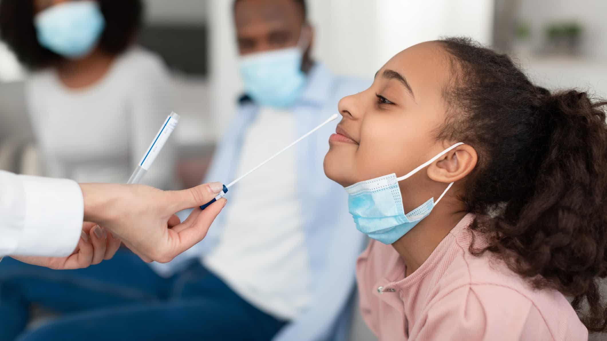 a young girl smiles as she is about to get a nasal swab test from a medical practitioner while her masked parent looks on in the background.