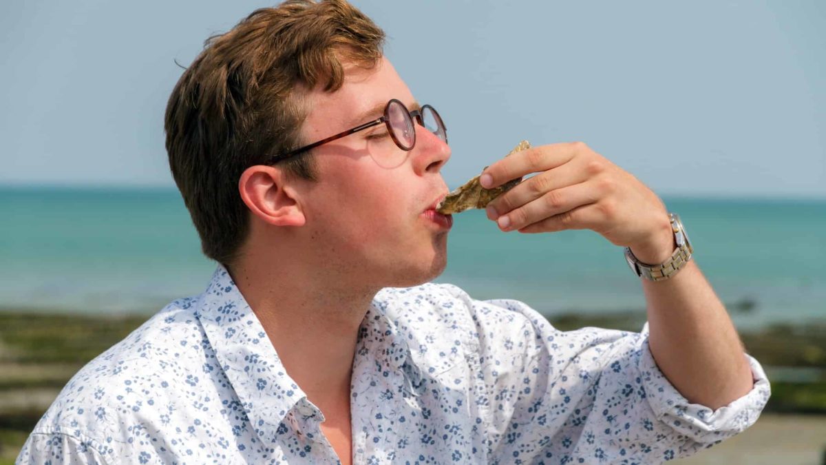 a man tips his head back to slurp down an oyster from its shell with a background of the ocean and a blue sky.