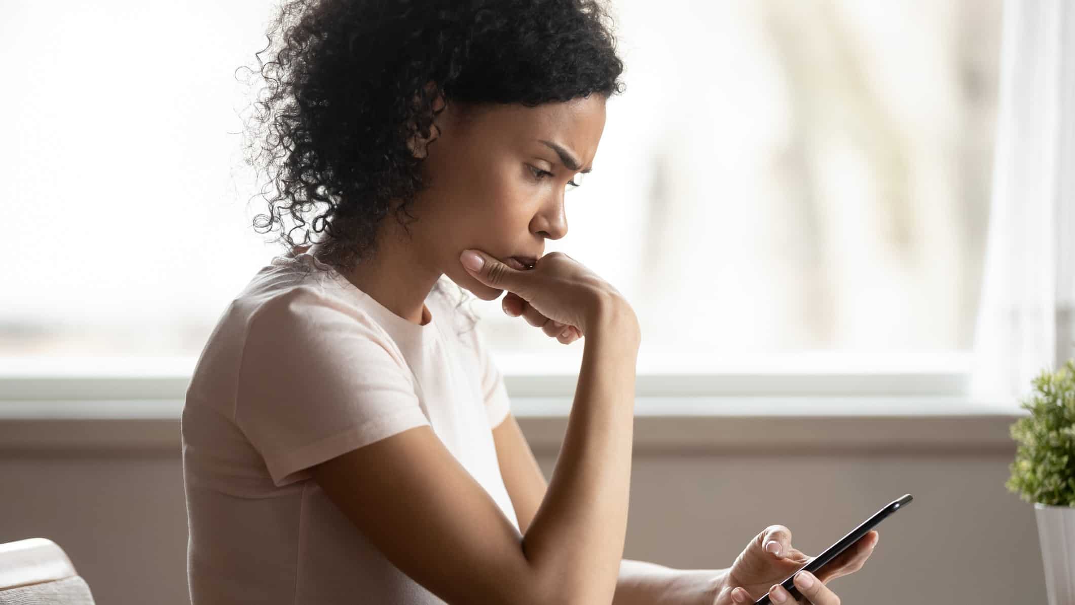 a woman looks down at her phone with a look of concern on her face and her hand held to her chin while she seriously digests the news she is receiving.