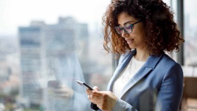 a woman in business wear looks at her phone against the window of a high rise space with a city landscape view of tall buildings outside.