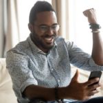 a man sits on his sofa loong at his phone and raises a fist to the air in happy celebration.
