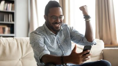 a man sits on his sofa loong at his phone and raises a fist to the air in happy celebration.