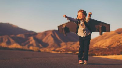 a young girl wearing a set of airplane wings stands on a tarmac with hands in the air and an excited look on her face as though she is about to take off.