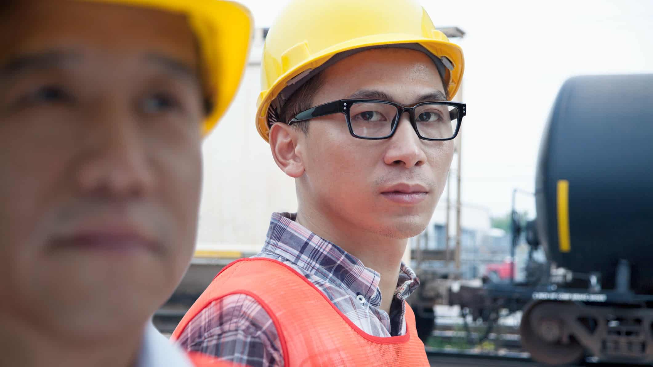 a man in a hard hat and high visibility vest gives a sombre look to the camera in the foreground of a rail freight yard.
