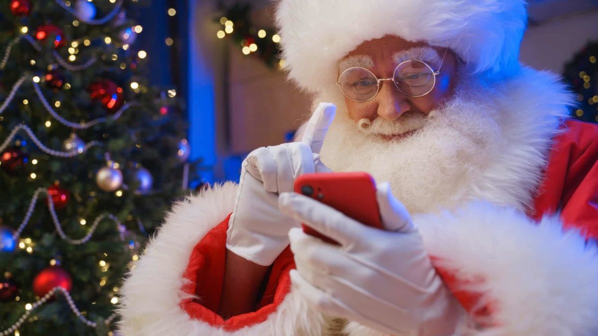 santa looks intently at his mobile phone with gloved finger raised and christmas tree in the background.