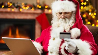 santa claus sits at a computer holding a card in his hand and looking at it with a hearth in the background next to a lit-up christmas tree.