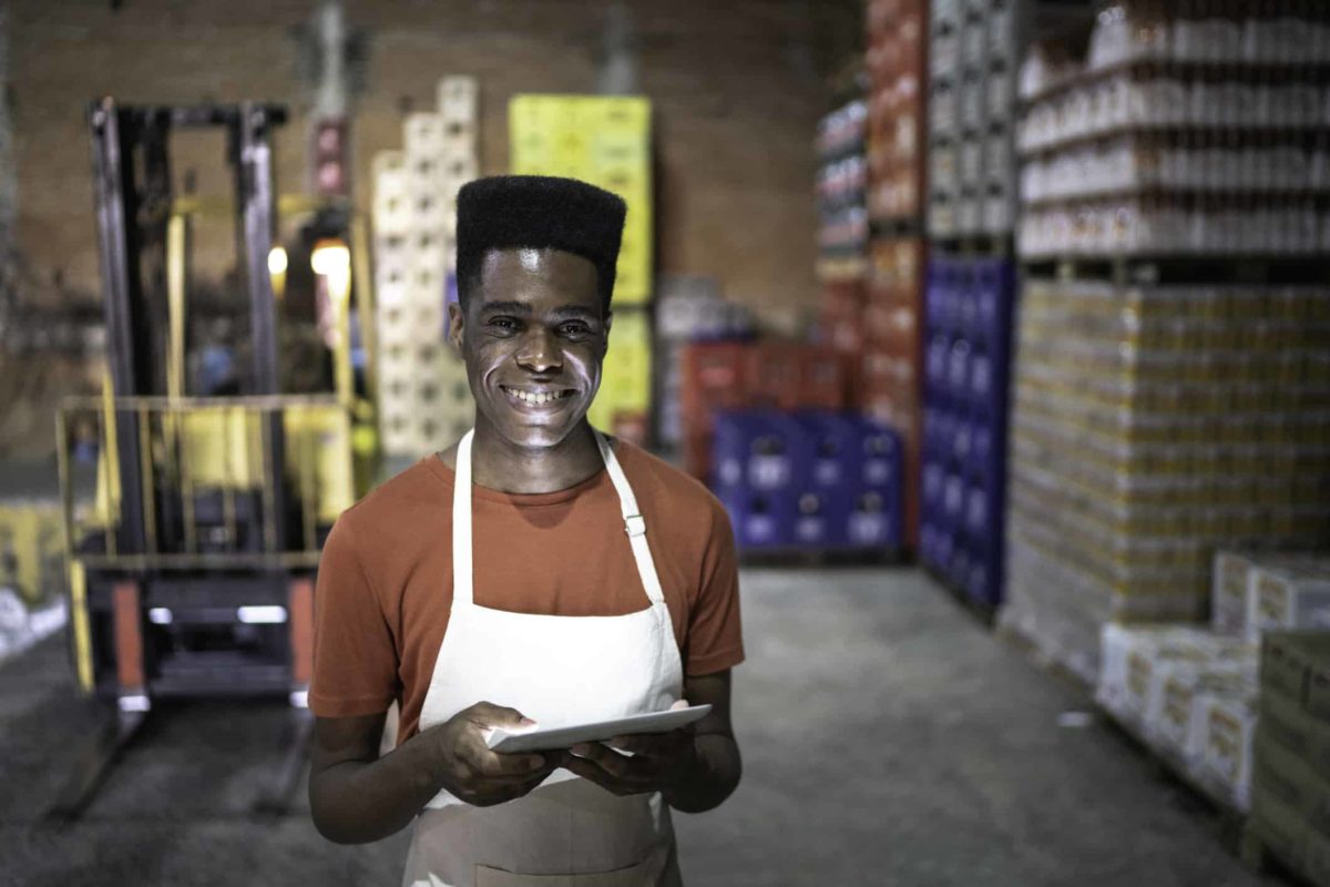 a smiling employee of an online shopping facility stands with a tablet in hand in front of a warehouse of goods and a vehicle to transport them.