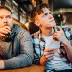 Two men in a bar looking uncertain as they hold a betting slip and watch TV.