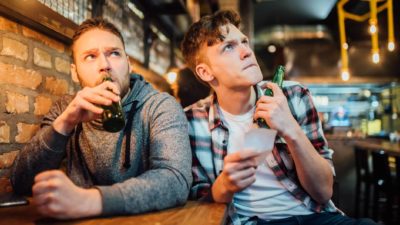 Two men in a bar looking uncertain as they hold a betting slip and watch TV.