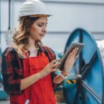 A woman wearing a hard hat and high visibility vest checks her device in front of a large spool of steel cable.