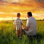 A man and his small son crouch in a green field under a beautiful sunset sky looking at renewable, wind generators for energy production.