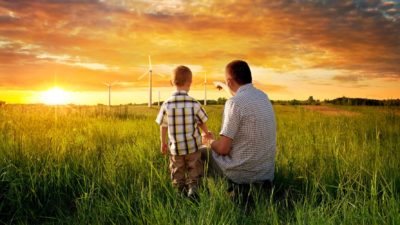 a man and his small son crouch in a green field under a beautiful sunset sky looking at renewable, wind generators for energy production.