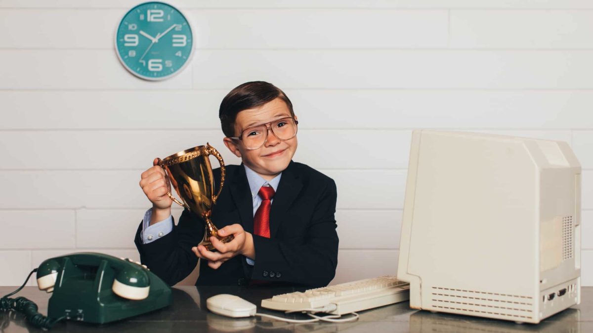 A young boy dressed in a suit and glasses that are too big for him sits at a desk and holds up a trophy representing the top 10 ASX shares today