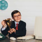 A young boy dressed in a suit and glasses that are too big for him sits at a desk and holds up a trophy representing the top 10 ASX shares today