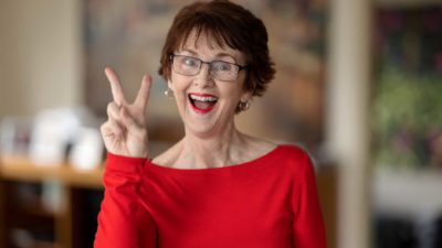 a middle-aged woman holds up two fingers with a wide mouthed smile on her face and wide open eyes.